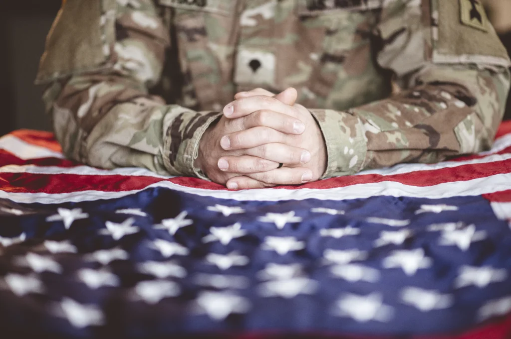 A veteran in military uniform clasps their hands on an American flag draped over a table.